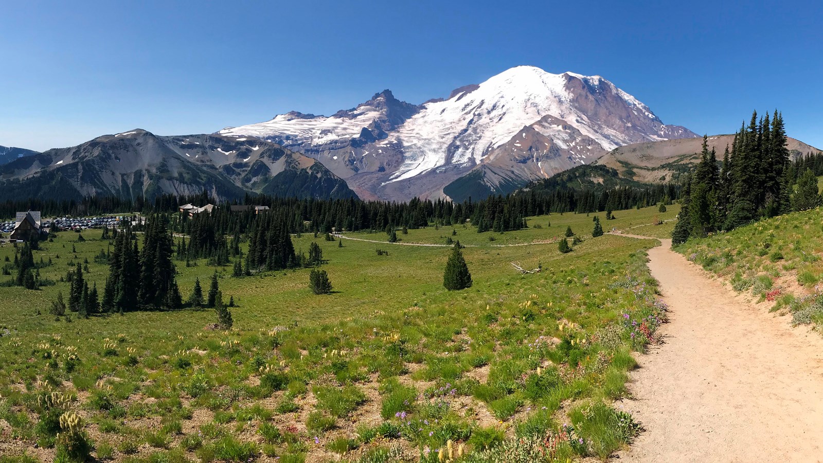 A gravel trail cuts down through a meadow towards a cluster of buildings on the slopes of a mountain